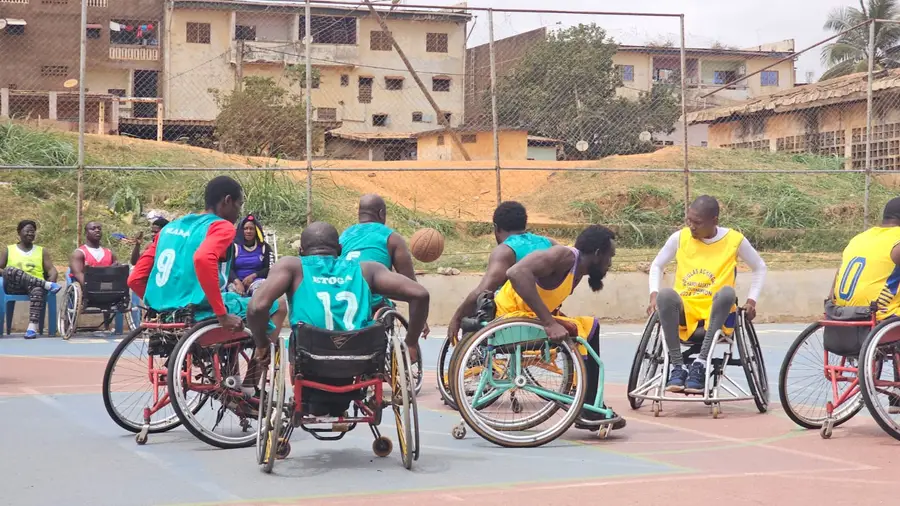 Match en plein air : joueurs en maillots verts et jaunes se disputent le ballon en fauteuil sur un terrain extérieur.