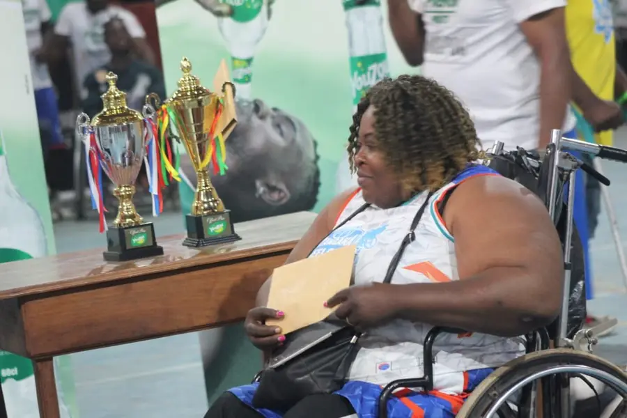 Une femme en fauteuil sourit devant la table des trophées, une enveloppe à la main.